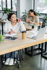 businesswoman in wheelchair talking on mobile phone near laptop and blurred african american colleague