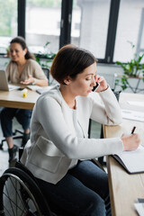 businesswoman with physical disability writing in notebook during conversation on smartphone in office