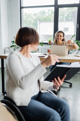 blurred businesswoman with disability pointing at notebook near african american colleague working...