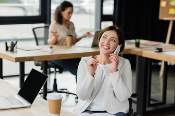 cheerful woman with disability talking on smartphone near laptop with blank screen and blurred african american colleague