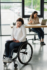 businesswoman in wheelchair talking on mobile phone and writing in notebook near blurred african american colleague