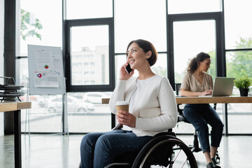 smiling businesswoman in wheelchair holding coffee to go and talking on cellphone near blurred african american colleague