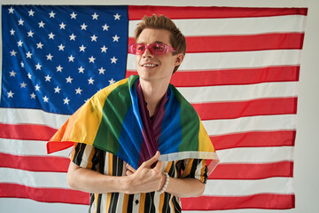 Smiling man is holding rainbow LGBT color flag at his shoulders on USA background