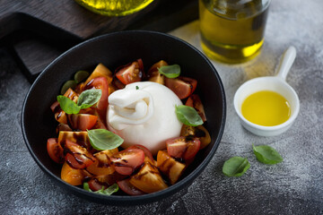 Salad with burrata cheese, cherry tomatoes, balsamic and green basil served in a black bowl, studio shot on a dark-grey stone background