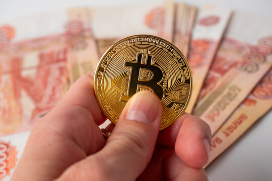a man's hand holds a gold bitcoin. In the background, five thousand Russian ruble bills. Cryptocurrency Concept