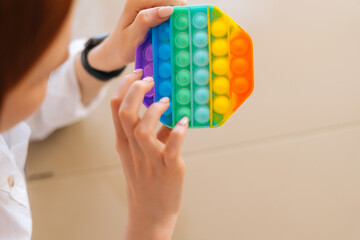 Close-up high-angle view of of unrecognizable young woman playing with rainbow pop-it fidget toy sitting at table. Closeup of female pushing colorful iridescent soft silicone bubbles sitting at desk.