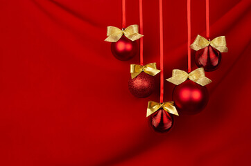 Shimmer red balls with gold bows hang on ribbons in row on deep scarlet silky smooth wavy wall, closeup, copy space. Luxury New year background for design of poster, flyer, card, brochure.