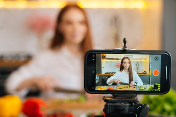 Display of camera recording video blog for food blogger woman cutting vegetable in modern kitchen talking about healthy vegan eating. Influencer vlogger girl live streaming nutrition masterclass.