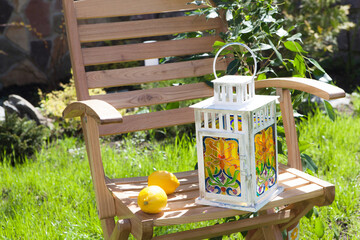 A homemade candlelight with multicolored stained glass stands on a white wooden chair on a green lawn in the summer. Two lemons lie side by side - obrazy, fototapety, plakaty