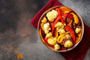Baked spicy vegetables in a bowl on a dark background top view. Pepper, sweet potato, cauliflower and onion baked with cumin, turmeric, curry and ginger. Indian food, vegan food. Copy space