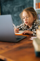 Close-up vertical shot of cute focused pupil child school girl doing homework writing notes with pen in paper notebook and looking at laptop computer. Concept of online e-learning education .