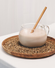 Non diary buckwheat milk. Vegan food composition with glass cup of plant milk with wooden straw on a wooden plate with buckwheat groats on a white table. Veggies healthy milk product