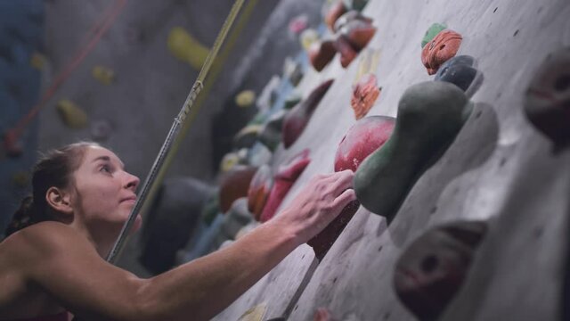Sporty girl rock climber is engaged on an artificial wall in the gym. Athletic Woman Grasping stones and ledges. Climbs up using a safety rope and a carabiner. Bouldering in the city. Slow motion.