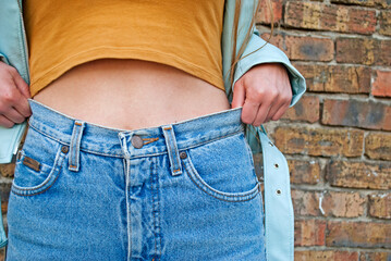 Hipster girl wearing jeans and leather jacket posing against a street wall, urban clothing style. Pull up your pants. 