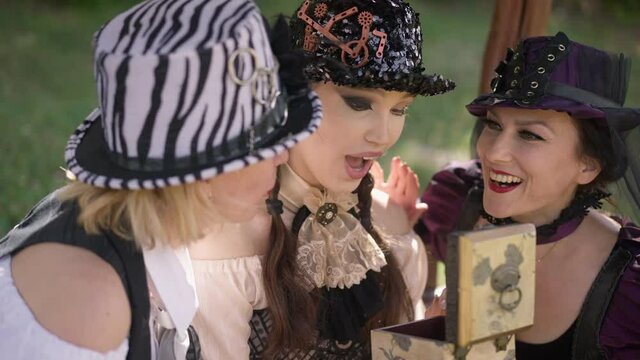 Surprised young woman opening gift box with amazed facial expression sitting in pavilion with friends. Happy carefree Caucasian women in steampunk outfit outdoors in sunshine. Slow motion