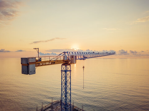 Close-up view from a drone of a construction tower crane against the background of a fantastic, beautiful sunset on the sea with a copy of the space