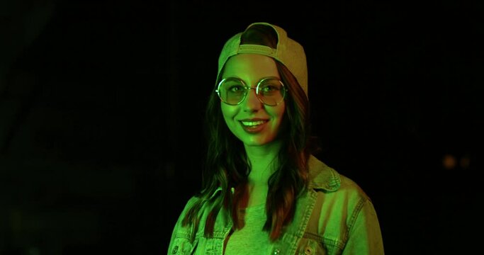 Portrait view of the cool stylish young caucasian hipster girl with long hair, sunglasses and cap posing to the camera at the neon light outside