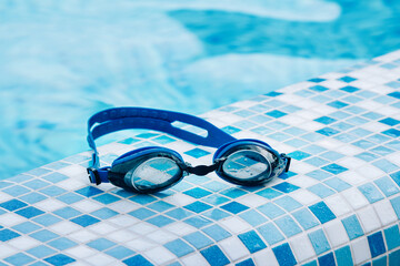 Blue professional swimming goggles with water drops on yellow lenses on a blue and white tiles of pool floor.