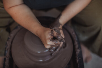 A middle-aged plus size woman in a pottery apron creates a clay vase on a potter's wheel in a pottery workshop