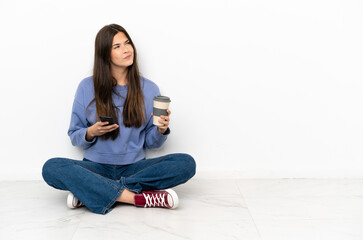 Young woman sitting on the floor holding coffee to take away and a mobile while thinking something