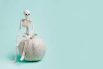 Human skeleton sitting on the fall gourde fresh pumpkin against pastel green background. Creative...