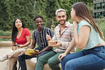 Group of young cosmopolitan multi-cultural friends students or colleagues enjoying conversation while eating take-out street food in the city park. Focus on the African guy