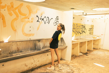 Young urban teenager girl showing tongue and standing up pretending to urinate as a man inside an abandoned public toilet with graffiti drawings on the wall. Youth freedom feeling.