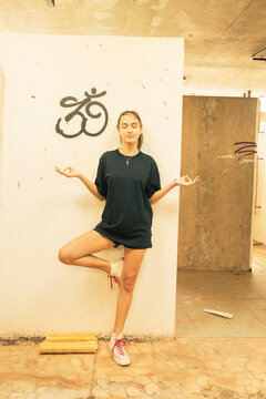 Young urban teenager girl standing up on one leg making a four and hands facing up as meditating inside an abandoned building with a graffiti drawing on the wall behind her. Youth freedom feeling.