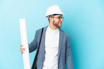 Young architect man with helmet and holding blueprints over isolated background looking side