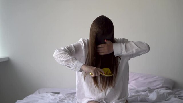 Serious girl 20s combs her hair with a hair styler while sitting on the bed. Taking care of the health and length of the comb. Prevention of hormonal imbalance in a woman's life.