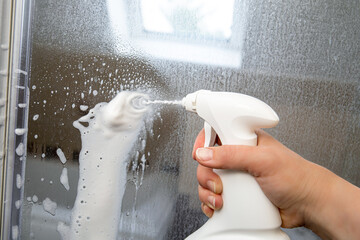 Person hand using limescale remover chemical foam. Descaling shower case glass door. Work in...