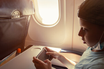 The boy looks at his mobile phone and listens to entertainment in flight on board the plane near the window.