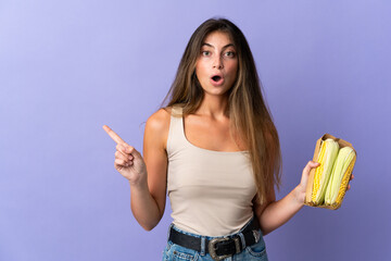 Young woman holding corn isolated on purple background surprised and pointing side