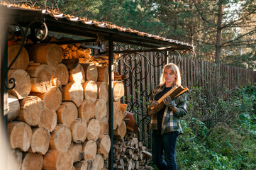 A beautiful blonde woman in a plaid shirt collects wood from a wood-burner. Everyday rural life.