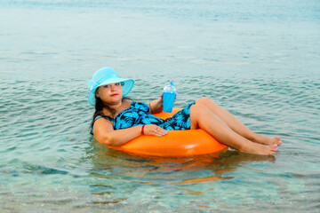 A woman in a straw hat and glasses in the sea in a swimming circle with a cocktail in her hand.