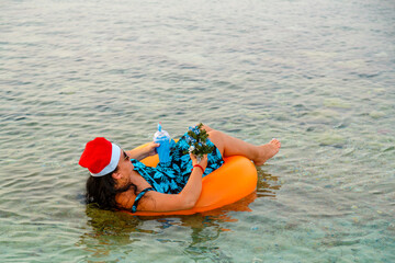 A woman in a swimming ring in a Santa Claus hat with a cocktail and a small Christmas tree in her hand floats in the sea. Shooting from the back.