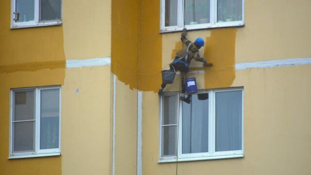 paint color high rise building on street on cables in helmets. facade worker with brush. safety construction with lift rope belt in city update. Reconstruction of residential town yellow color.
