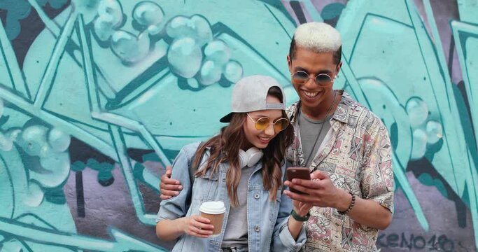 Cute and cheerful multiracial hipsters couple, boyfriend and girlfriend in stylish outfits looking the smartphone on the graffity wall outdoors while spending time together