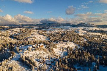Aerial winter landscape with small village houses between snow covered forest in cold mountains in the evening.
