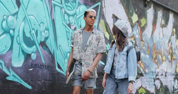 Street shot of the young diverse hipsters couple in love chatting with each other at the graffity wall outside and walking with skateboard in front of the camera