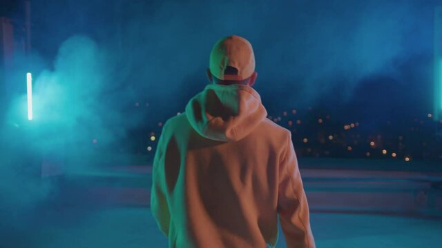 Back of hiphop stylish bboy dancing guy in yellow outfit walking outside on night stage rooftop and watching cityscape. Artisy performance. Nightlife.