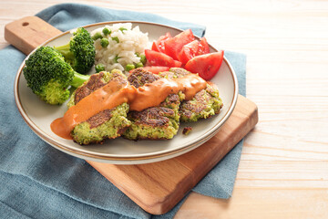 Vegetable patties from broccoli with tomato, rice and a hot sauce, vegetarian and vegan meal on a cutting board, blue napkin and light wooden table, copy space, selected focus