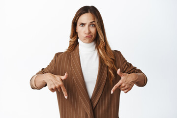 Skeptical female company ceo, grimacing and looking with disbelief, pointing fingers down at advertisement below, standing in brown suit against white background