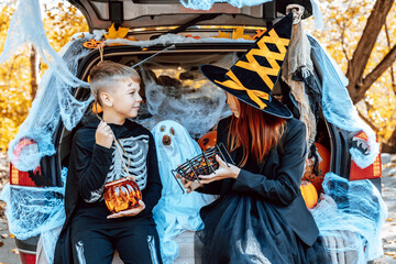 Obraz premium siblings boy in skeleton costume, teenage girl in witch costume and hat and cute poodle dog in ghost costume sits in trunk car decorated for Halloween with web, orange balloons and pumpkins, outdoor