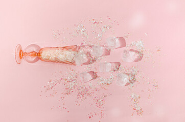 A pink Champagne glass filled with shiny tinsels on pink background. Ice cubes and tinsels around the glass. Creative New Year or Christmas celebration concept. Trendy visual trend backdrop. Flat lay.