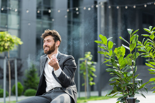 Caucasian bearded business man smoking cannabis outdoors sitting on city park bench on urban street background. Male employee Businessman. Office worker in suit Relieves stress a marijuana outside