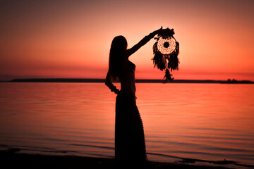 Silhouette of a woman with a dreamcatcher in her hand on a background of sunset over the lake