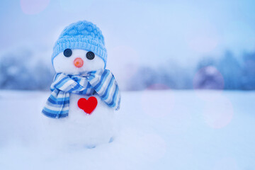 Little cute snowman with a red heart in a blue knitted hat and scarf on the snow on a winter day....