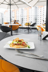 Croissant sandwich with bacon, tomatoes and lettuce salad on white plate and bright background. Continental style breakfast.