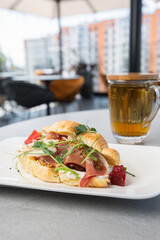 Croissant sandwich with prosciutto, berries, dijon mustard, cheese and cup of tea on white plate and bright background. Continental style breakfast.